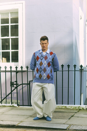 Man in patterned argyle sweater stands in front of building with wrought-iron fence in an outdoor setting, casual fashion shoot.