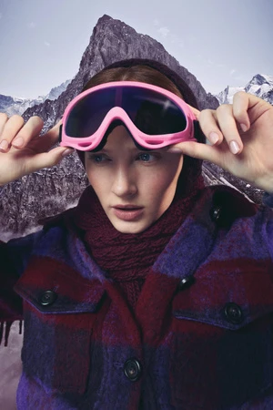 A woman wears pink ski goggles and a patterned jacket with a mountain backdrop, embodying winter fashion and outdoor style.