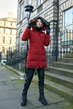 A woman in a red Canada Goose parka, black pants, and boots stands in an urban setting with historic buildings and a streetlight.