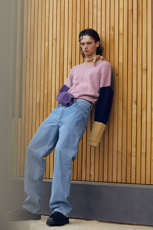 Young adult leans against wood slats wearing light wash jeans and pink color-blocked sweater with black shoes and a beige scarf.