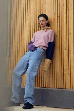 Young adult leans against wood slats wearing light wash jeans and pink color-blocked sweater with black shoes and a beige scarf.