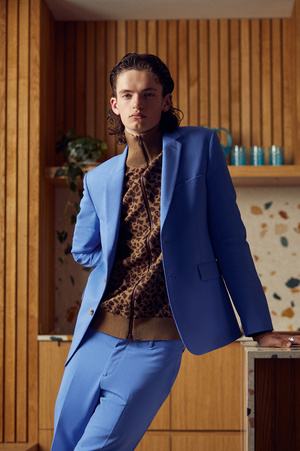 Stylish young man in a blue suit and animal print sweater strikes a pose indoors, leaning on a counter in a brightly lit scene.