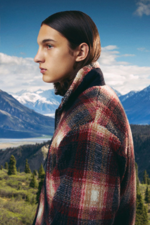 Profile portrait of a stylish young adult with slicked-back hair wearing a plaid coat in front of a mountain range backdrop.