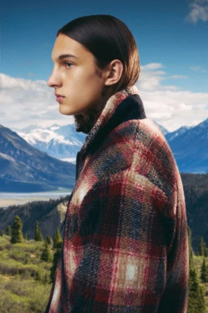 Profile portrait of a stylish young adult with slicked-back hair wearing a plaid coat in front of a mountain range backdrop.