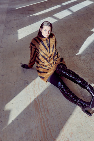 A woman poses on a light-colored floor, wearing a tiger-striped sweater and thigh-high black boots, illuminated by natural light and shadows.