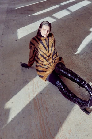 A woman poses on a light-colored floor, wearing a tiger-striped sweater and thigh-high black boots, illuminated by natural light and shadows.