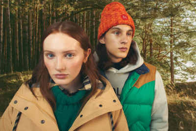 Two young adults stand in a forest setting, wearing casual outerwear in earthy tones. Close-up, eye-level, outdoor fashion shot.