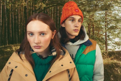 Two young adults stand in a forest setting, wearing casual outerwear in earthy tones. Close-up, eye-level, outdoor fashion shot.