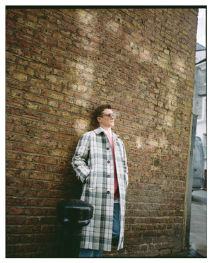 Adult male standing next to a brick wall wearing a plaid coat and sunglasses in an urban setting with a street and car in the background.