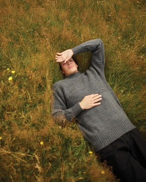 A man wearing a grey knitted sweater lying among tall wild grass and yellow wildflowers, photographed from above by Stew Bryden.