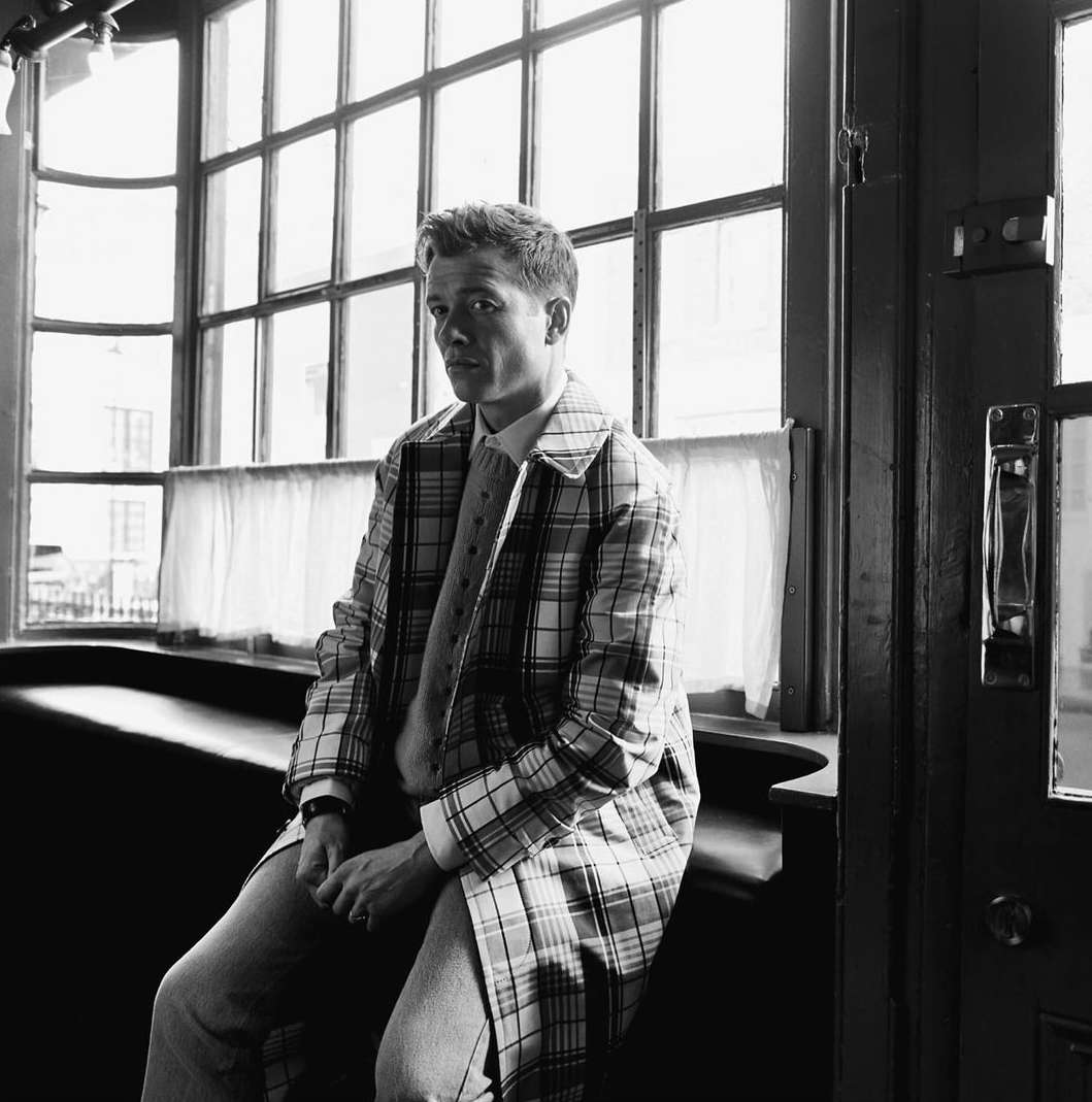 Monochrome portrait of a man in a plaid coat sitting in front of a window, wearing a tie and light-colored jeans.