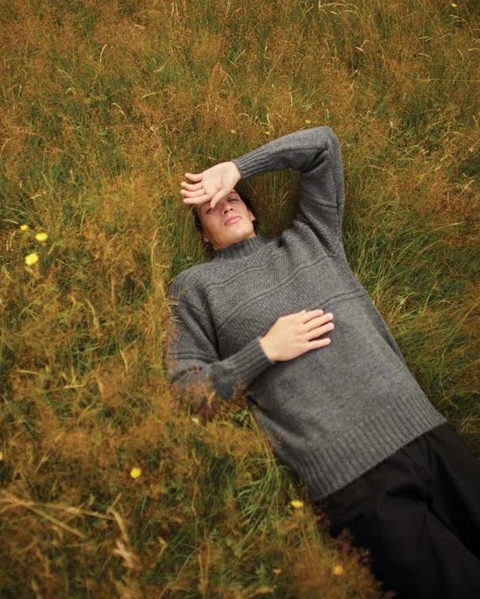 Man lying in a tall grassy meadow wearing a grey knit sweater and shading his eyes from the sun.