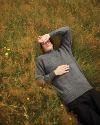 Man lying in a tall grassy meadow wearing a grey knit sweater and shading his eyes from the sun.