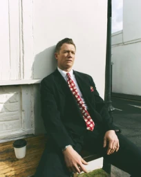 A man in a black suit and patterned red tie sitting outdoors against a white wall by Stew Bryden