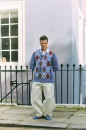 A man wearing an argyle sweater vest and white trousers stands outside against a pale purple building.