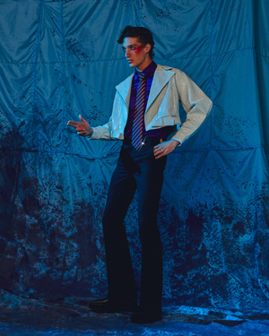A man in a cropped white jacket, purple shirt, striped tie, and dark trousers posing against a crinkled blue backdrop.