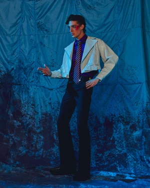 A man in a cropped white jacket, purple shirt, striped tie, and dark trousers posing against a crinkled blue backdrop.