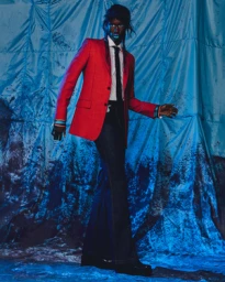 Model wearing a vibrant red blazer, white shirt, black tie, and dark trousers against a crinkled blue fabric backdrop.
