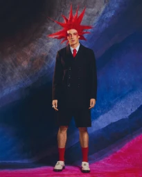 Man with vibrant red spiky hair wearing a black suit jacket, shorts, white shirt, red tie, and red socks against a painted backdrop.