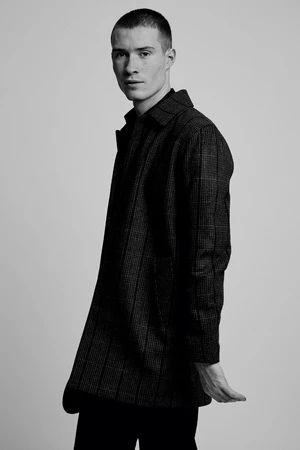 Monochrome portrait of a young man wearing a patterned coat against a seamless white background.
