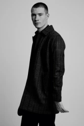 Monochrome portrait of a young man wearing a patterned coat against a seamless white background.