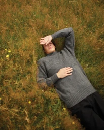 A man wearing a grey knitted sweater lying among tall wild grass and yellow wildflowers, photographed from above by Stew Bryden.
