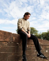 Young man sitting on a brick wall wearing a textured beige sweater, dark trousers, and black sneakers against a cloudy sky