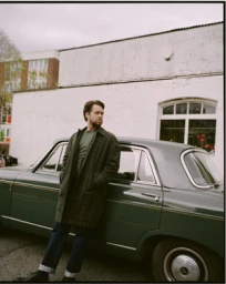 Man wearing a plaid coat leaning against a vintage green car in front of a white building exterior
