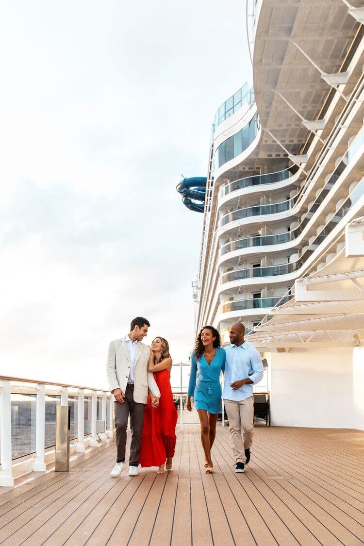 Two couples stroll on a cruise ship deck. One woman wears a red dress and the other wears a blue dress. The ship is white.