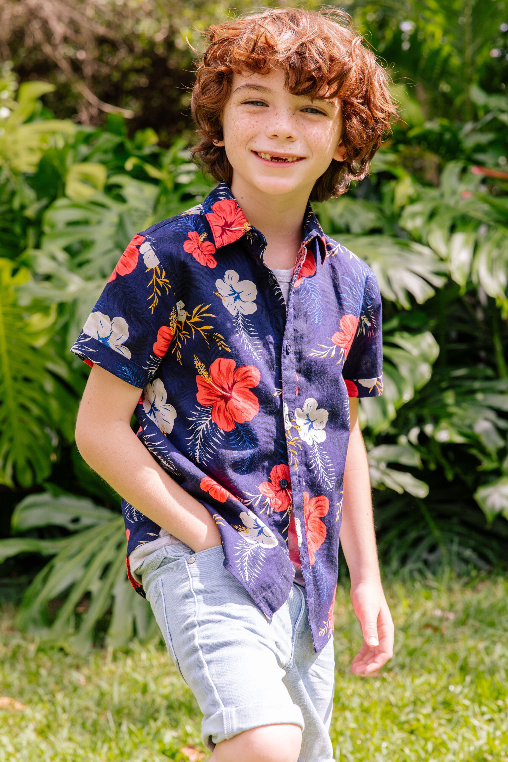 Smiling child with red curly hair poses outdoors in a vibrant floral shirt and denim shorts, surrounded by tropical plants and grass.