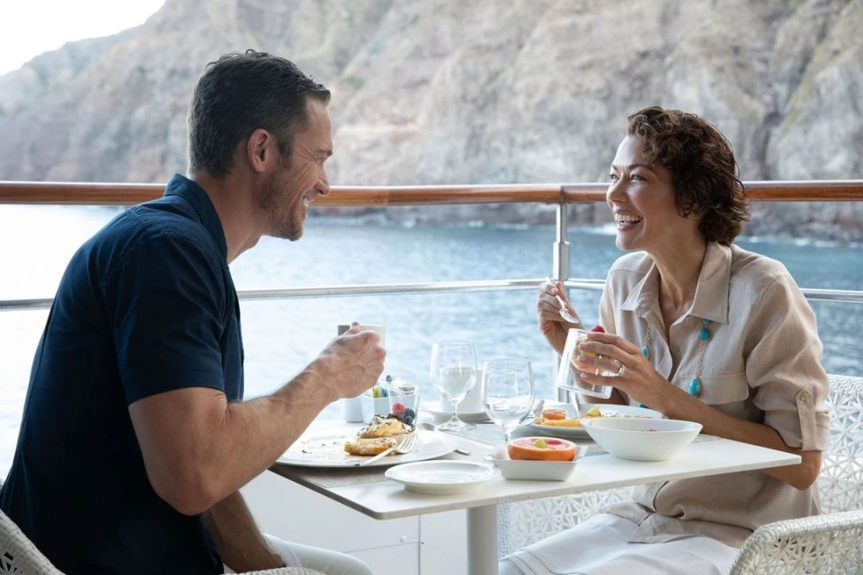 A smiling man and woman sit at a table outside, enjoying breakfast with a view of the sea and cliffs in the background.
