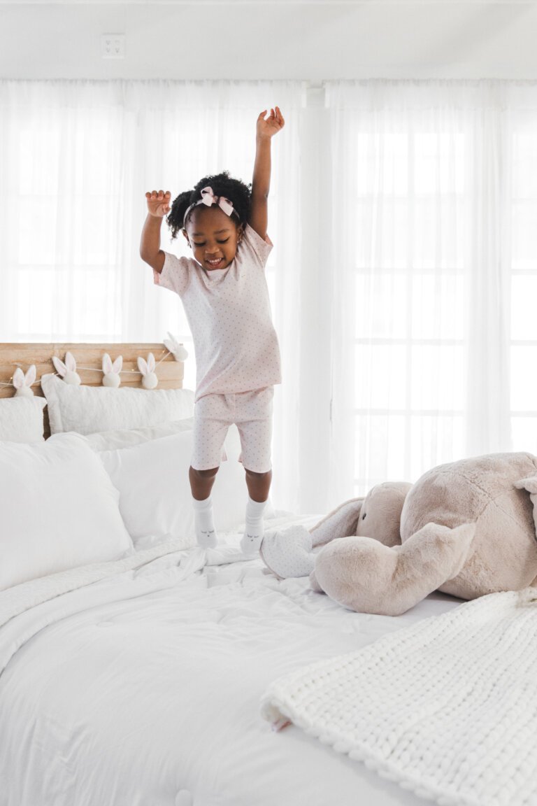 Young girl in pink pajamas jumping on a white bed with a stuffed rabbit toy, in a bright, naturally lit bedroom with white walls and bedding.