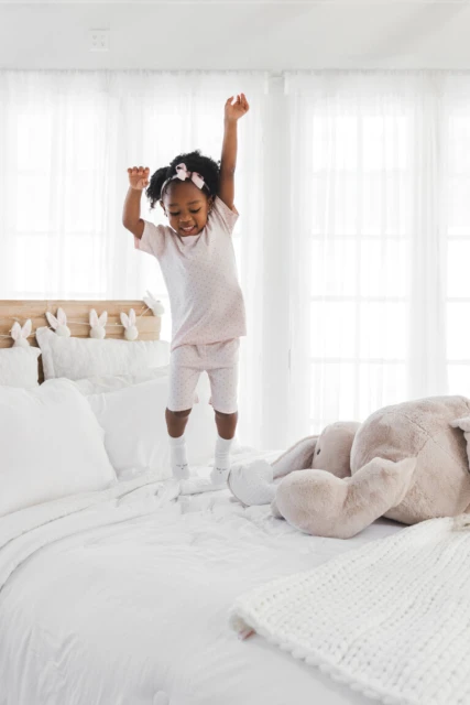 Young girl in pink pajamas jumping on a white bed with a stuffed rabbit toy, in a bright, naturally lit bedroom with white walls and bedding.
