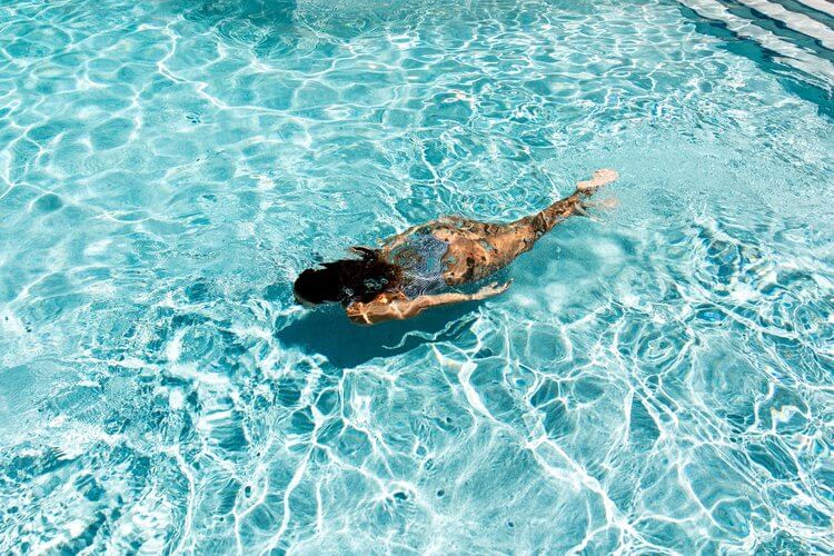 Woman swims in a turquoise swimming pool with sunlight creating patterns on the water, creating a sense of relaxation and summertime fun.
