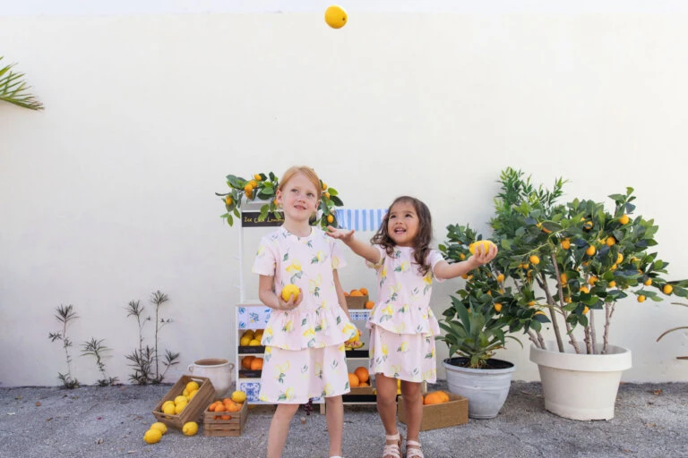 Two young girls in matching dresses play with lemons near a lemonade stand and potted citrus trees in a brightly lit outdoor scene.