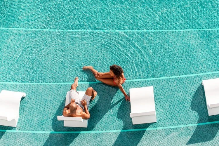 Aerial view of a man and woman relaxing on lounge chairs in a turquoise swimming pool with sunlight reflecting off the water.