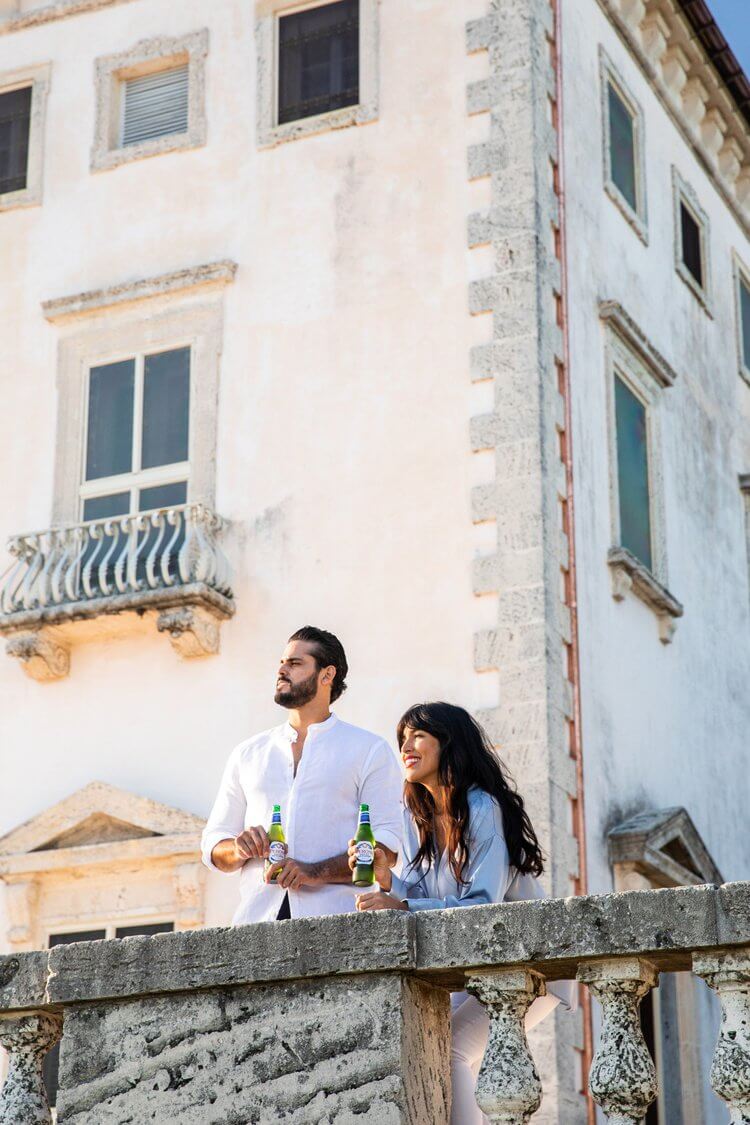 Adult couple, an adult male and adult female, standing on a balcony overlooking the city, holding beer bottles, on a sunny day.
