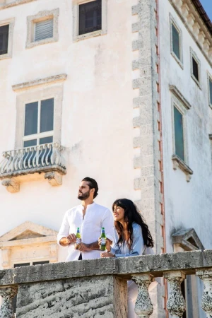 Adult couple, an adult male and adult female, standing on a balcony overlooking the city, holding beer bottles, on a sunny day.
