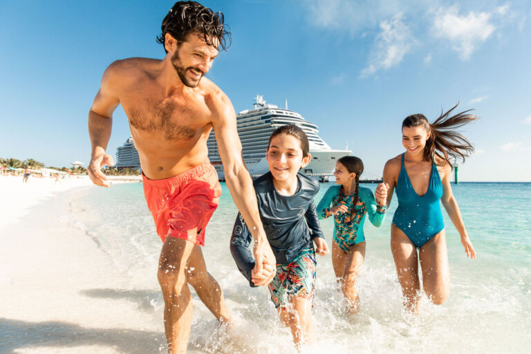 A family of four runs along a white sand beach toward the viewer with a large cruise ship in the background on a sunny day.