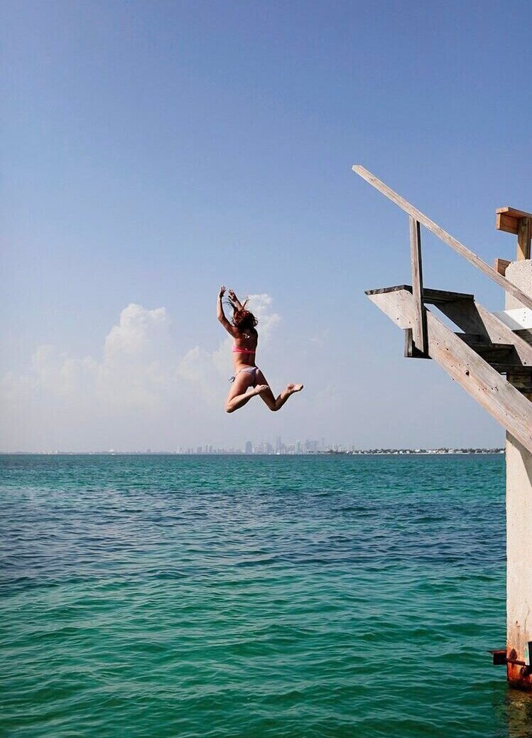 A woman in a swimsuit jumps from a wooden dock into turquoise ocean water under a clear blue sky with a cityscape on the horizon.