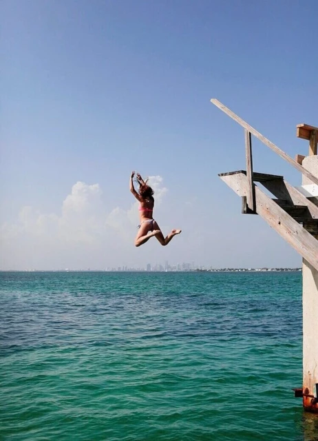 A woman in a swimsuit jumps from a wooden dock into turquoise ocean water under a clear blue sky with a cityscape on the horizon.