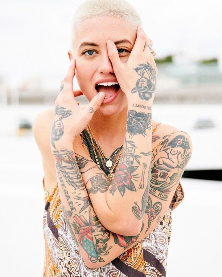 Tattooed woman with short blonde hair and patterned top covering her eye and sticking her tongue out in a studio shot.
