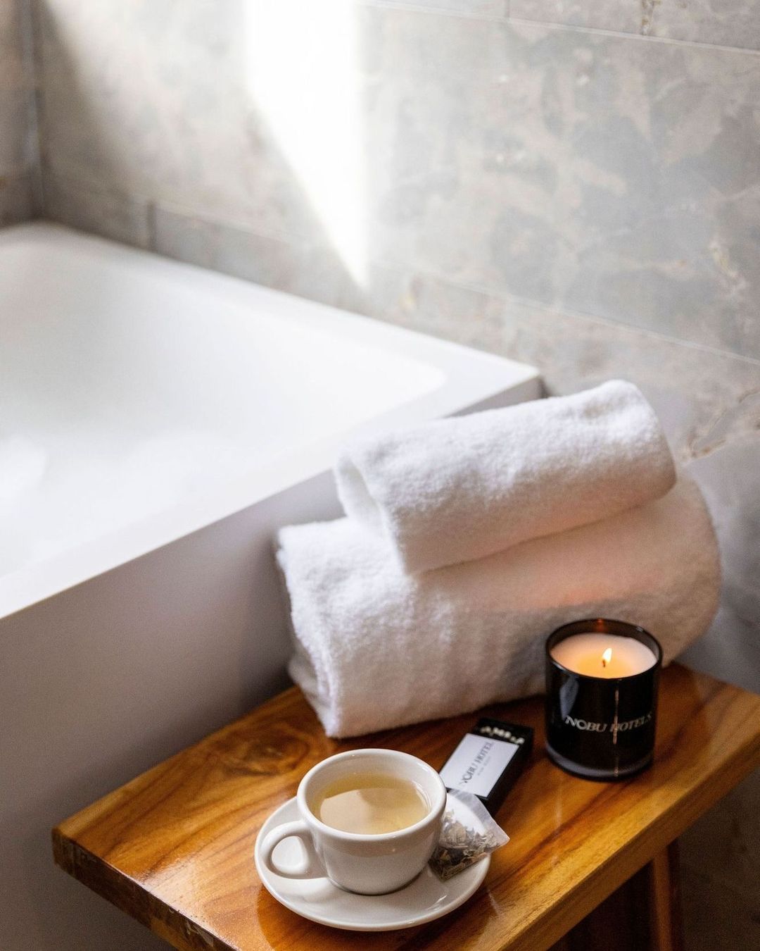A cup of tea, towels, and a candle sit on a wooden stool in a modern bathroom with marble walls, creating a spa-like atmosphere.