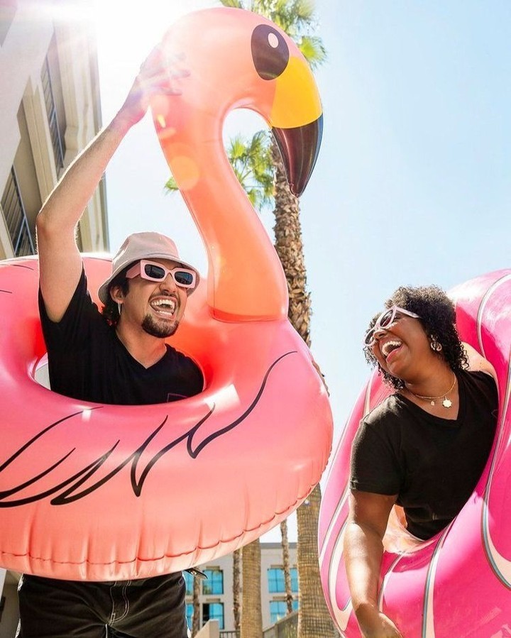 Two joyful people, a man and a woman, holding inflatable flamingo and inner tube at an outdoor pool, smiling with summer vibes.
