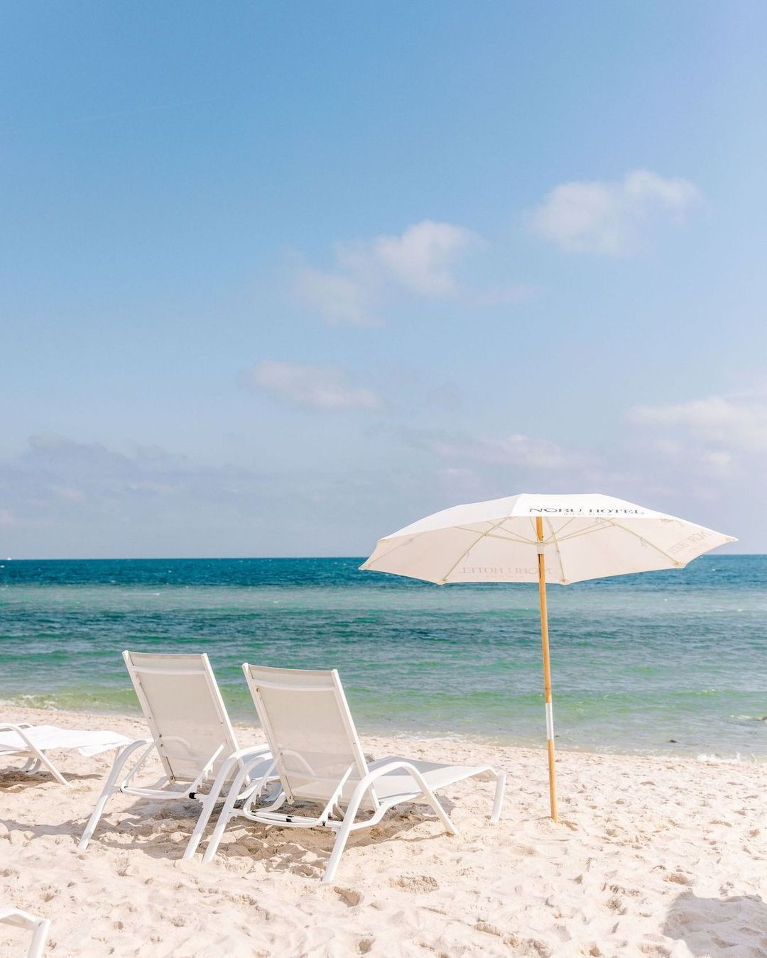 Two white lounge chairs and an open parasol on a sandy beach overlooking a calm blue ocean under a clear sky.