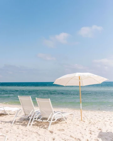Two white lounge chairs and an open parasol on a sandy beach overlooking a calm blue ocean under a clear sky.