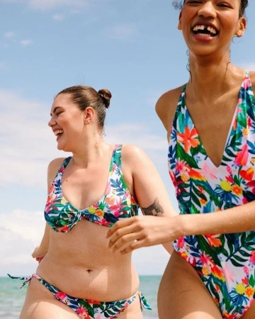 Two smiling women in floral swimsuits laughing on a sunny beach outdoors