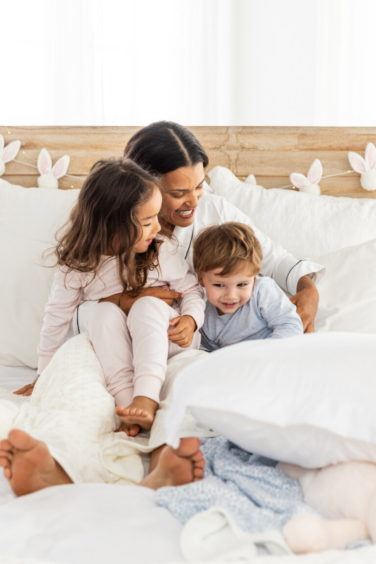 A mother is sitting on a bed with two children, a girl and a boy. All three are wearing pajamas and smiling in a light and airy setting.