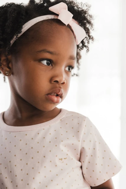 Close-up of a young African American girl with curly hair, wearing a pink polka-dot shirt and a pink headband with a bow, looking away.
