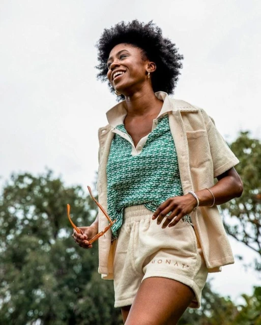 A smiling Black woman stands outdoors in a patterned shirt and beige shorts suit, holding sunglasses, with trees in the background.